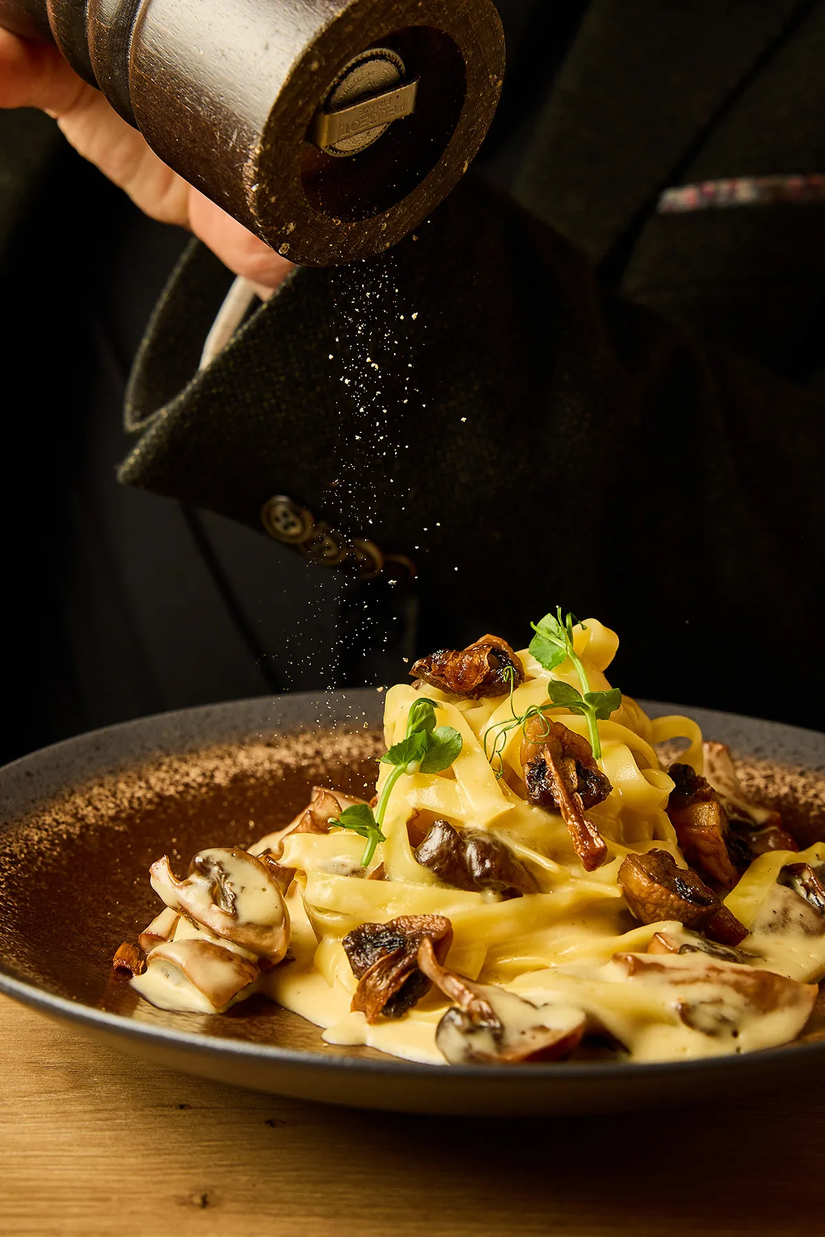 Main tenant un moulin à poivre déposant du poivre sur un plat de tagliatelles à la crème de truffe, champignons & parmesan, servies dans le restaurant In Casa à Levallois-Perret